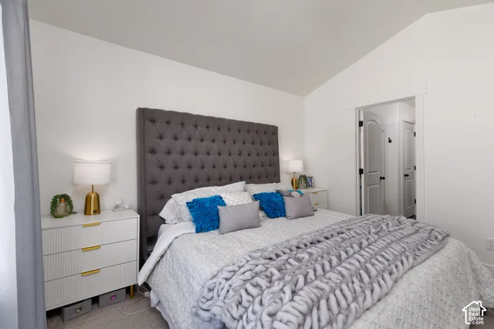 Vaulted ceiling, white walls, gray upholstered headboard, white side table with gold hardware, and white trim around the doorway