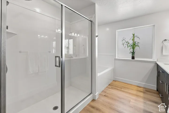 Glass-enclosed shower with a built-in shelf, wood-finish flooring, white soaking tub, dark-finish vanity with countertop, and frosted window