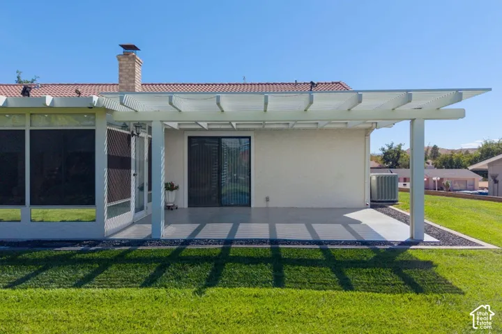 Expansive backyard featuring a green lawn, concrete patio, a white slatted pergola, and a stucco exterior with a sliding glass door