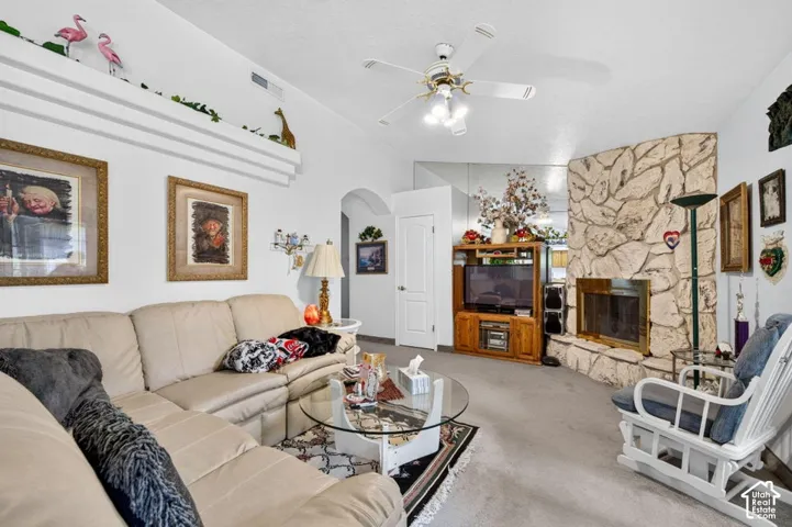 Built-in stone fireplace with brass-toned insert, wall-mounted shelving, arched doorway, ceiling fan with integrated lighting, and carpeted flooring