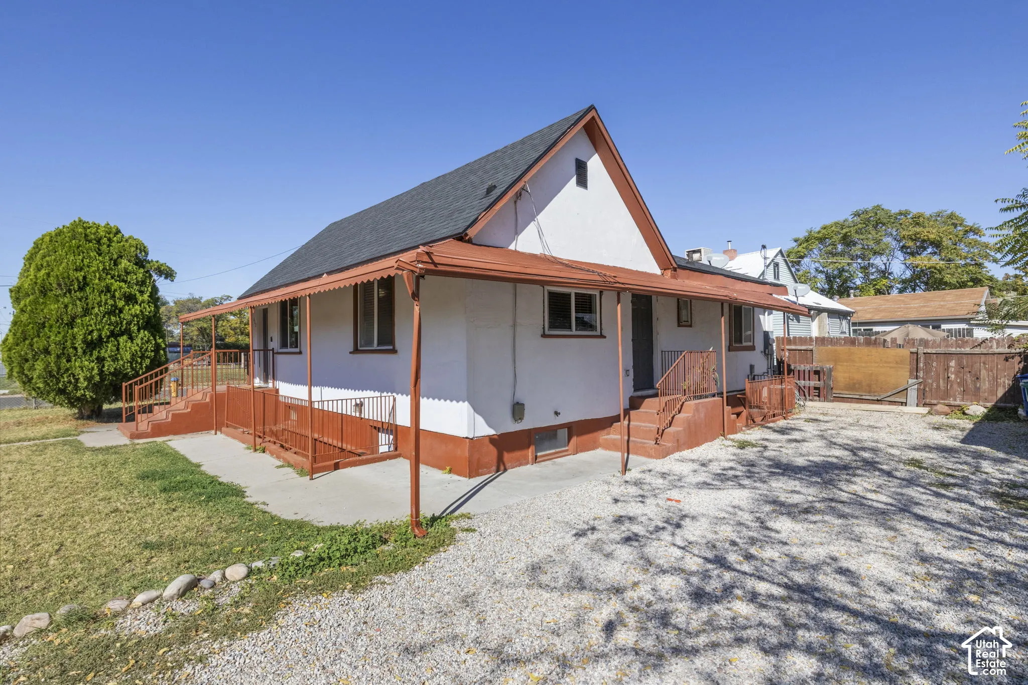 White stucco exterior with orange trim, covered porch, multiple entry points, gravel driveway, and a fenced yard