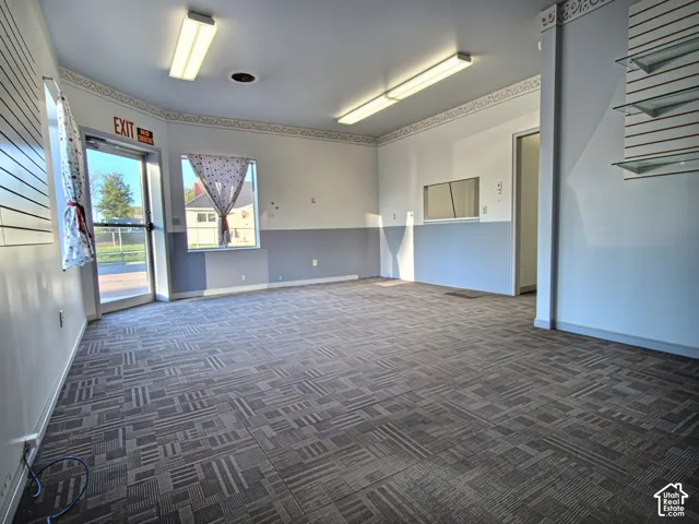 Spacious room featuring patterned carpet tiles, ample natural light from multiple windows, fluorescent overhead lighting, and built-in shelving with glass accents