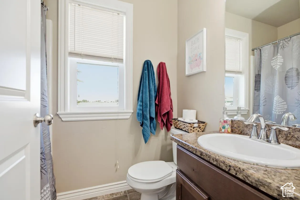 Bathroom featuring a single window, a toilet, an integrated sink with a chrome faucet, and a vanity with a stone-finish countertop