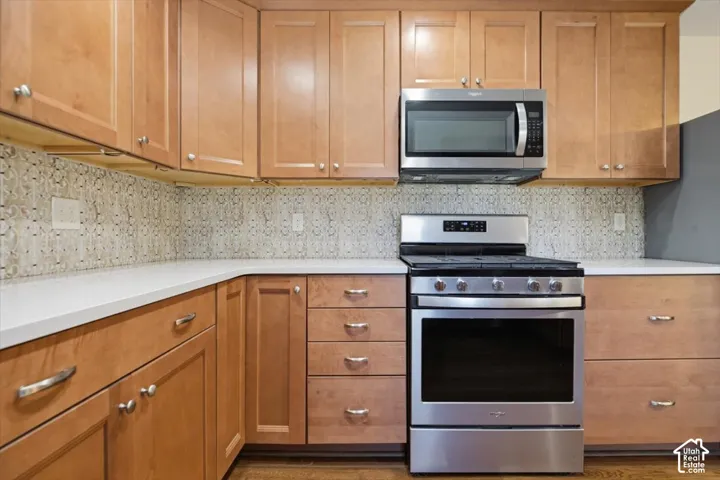 Wood-finish cabinetry, stainless steel range and microwave, light-toned countertops, and mosaic tile backsplash