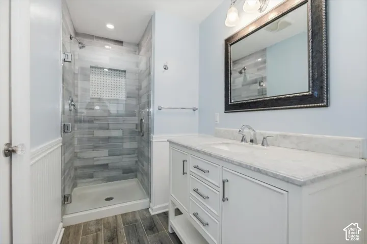 Glass-enclosed shower with tiled surround, single vanity with white cabinetry, stone countertop, and wood-finish flooring