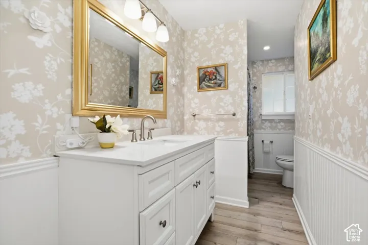 White vanity with integrated sink, gold-framed mirror, three-light fixture, wood-finish flooring, and white beadboard paneling