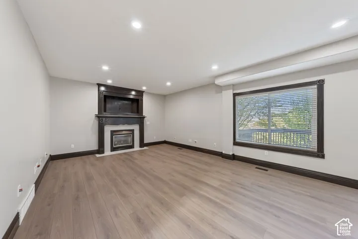 Recessed lighting illuminating wood-finish flooring, a decorative fireplace with an ornate wood mantel, and a large window with horizontal blinds