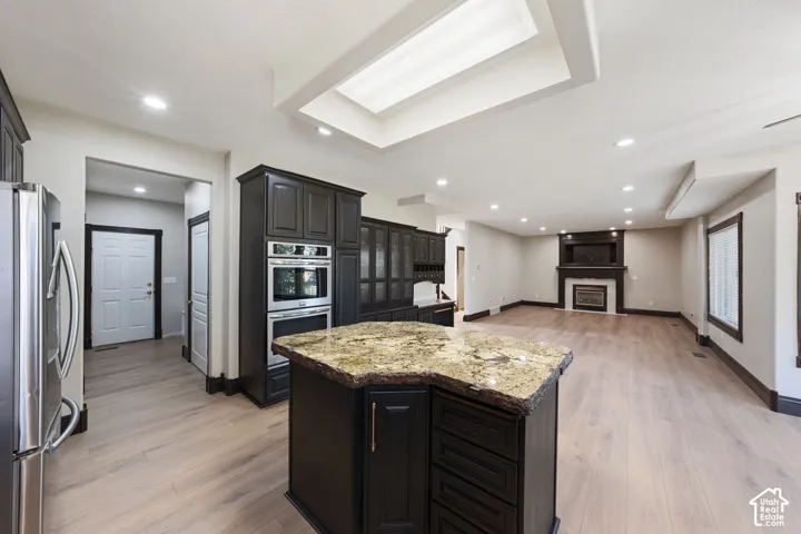 Chef's kitchen featuring a large center island with warm-toned stone countertop, dark cabinetry with glass-front uppers, stainless steel appliances including double wall ovens, and wide-plank wood-finish flooring throughout