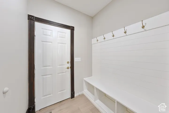 White paneled door with dark wood-finish trim, white shiplap wall with brass-finish hooks, built-in bench with cubby storage, and light wood-finish flooring