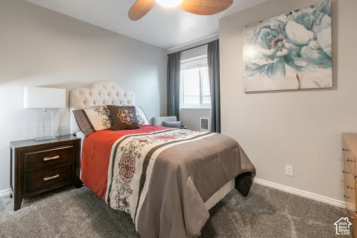 Tufted upholstered headboard, wood-finish nightstand with metal pull handles, window with dark drapes, ceiling fan with wood-finish blades, and neutral-tone carpeting