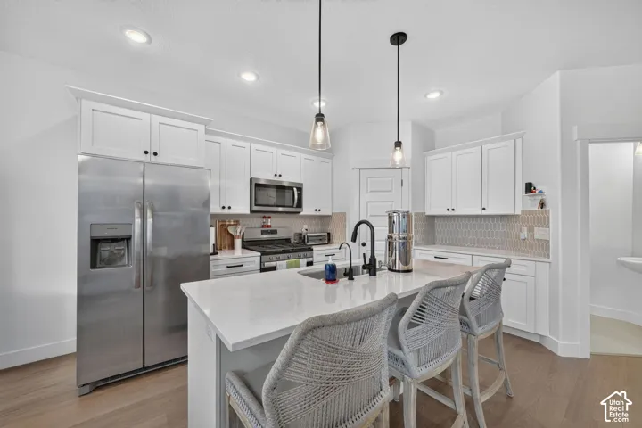 Contemporary kitchen featuring white shaker cabinetry, stainless steel appliances, a center island with a sink, pendant lighting, and wood-finish flooring