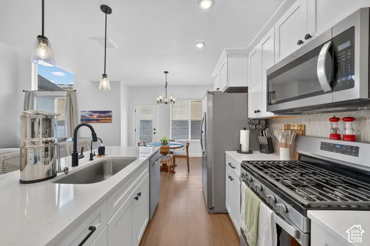 White cabinetry, stainless steel appliances, wood-finish flooring, island with integrated sink, and pendant lighting