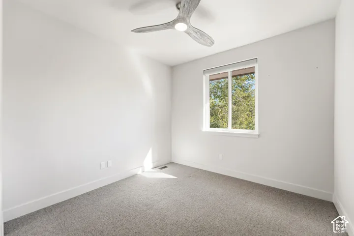 Crisp white walls, gray carpeting, a modern ceiling fan, and a large window with roller shades