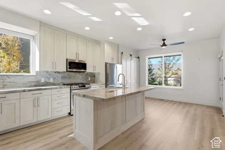 Modern kitchen featuring light wood-finish cabinetry, stainless steel appliances, a large island with a sink, stone countertops, and wood-finish flooring