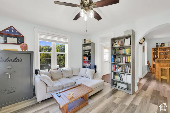 Wood-finish flooring, ceiling fan with light fixture, large window with white trim, arched doorway, and built-in shelving