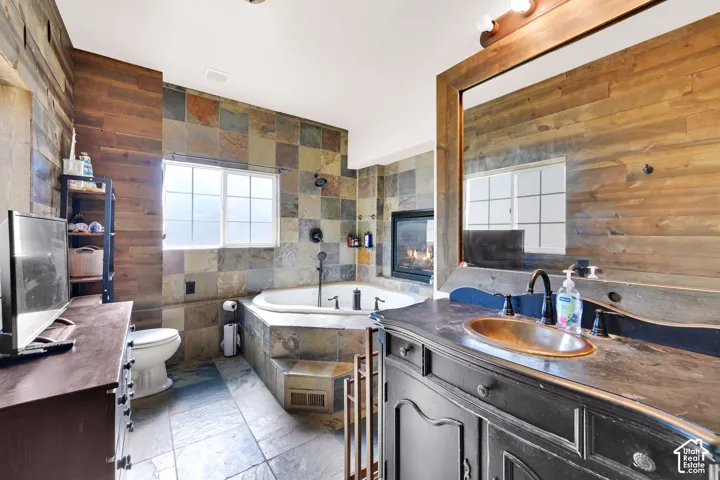 Corner soaking tub, stone tile flooring, wood panel wall accents, fireplace, and vanity with an integrated sink