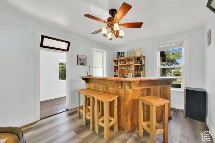 Reclaimed wood-finish bar with integrated shelving, wood-finish flooring, ceiling fan with lighting, and white-framed windows