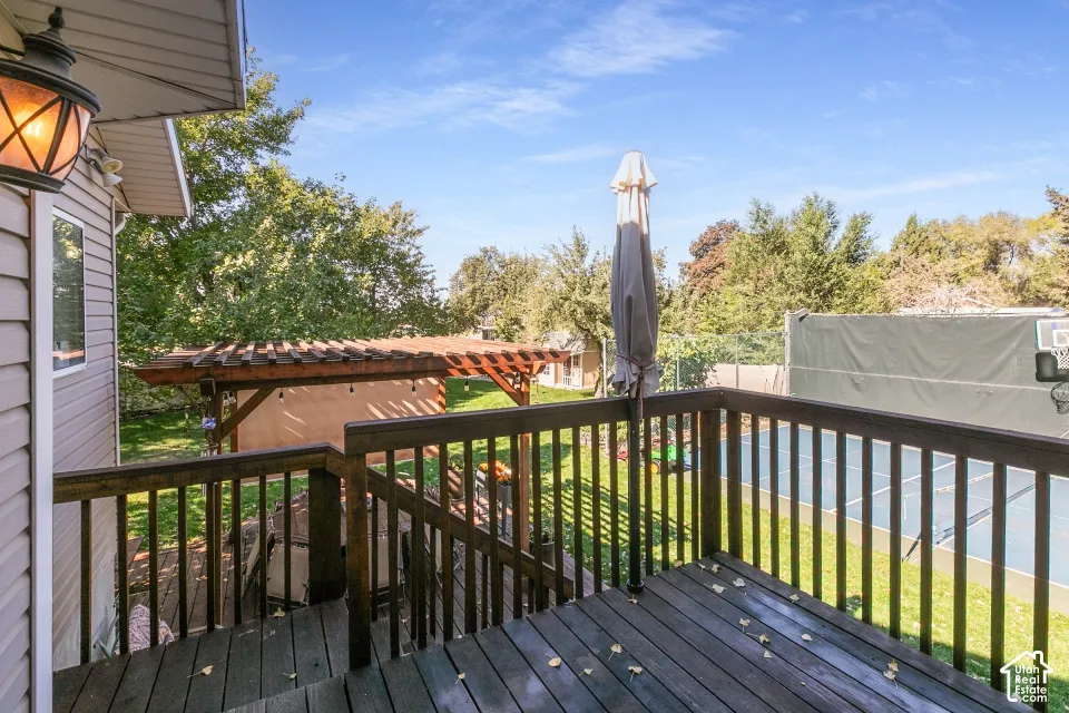 Wood-finish deck with railing, providing access to a pergola-covered structure and a paved court with a basketball hoop