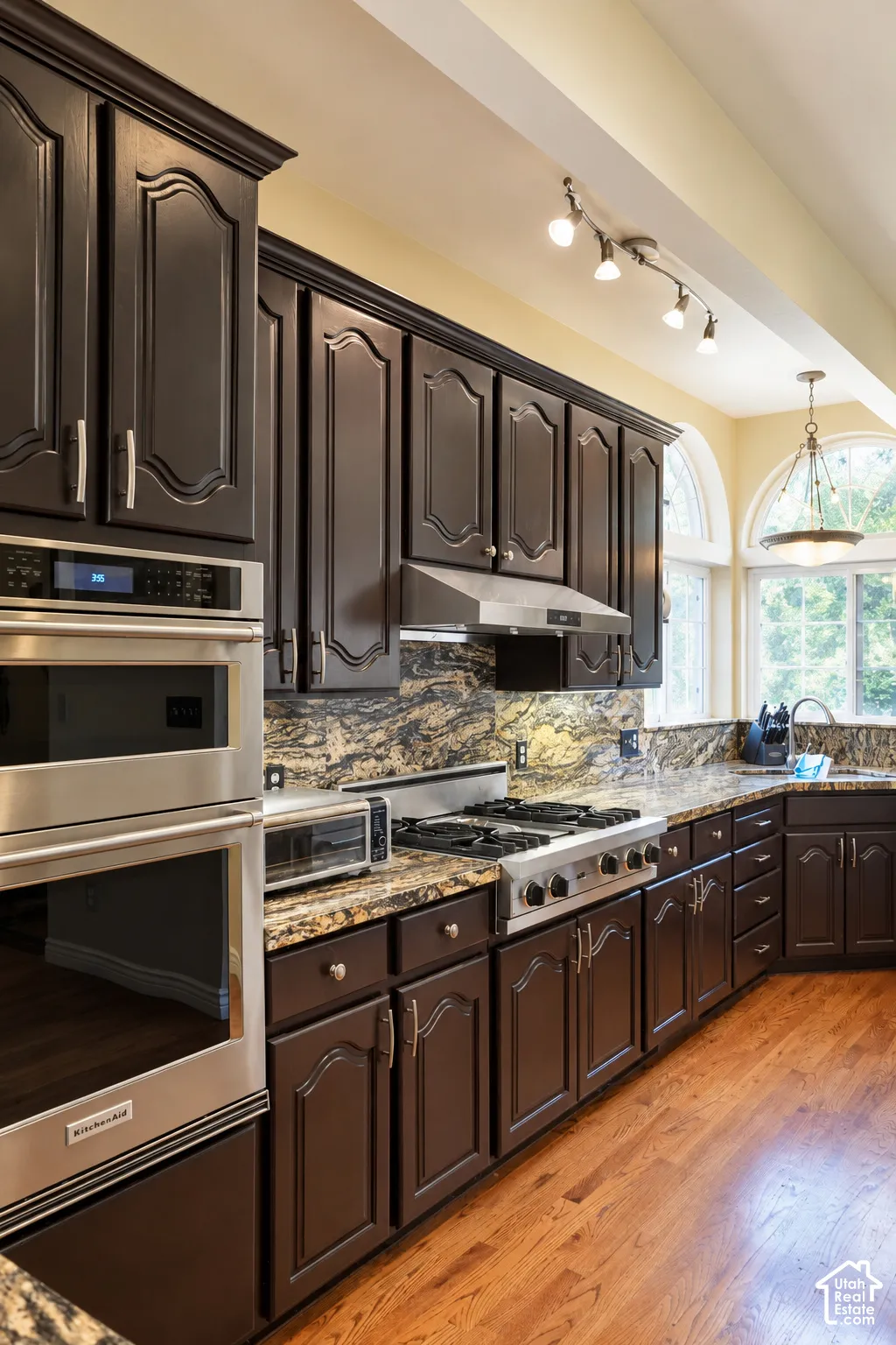 Dark wood cabinetry, stainless steel double ovens, gas range, patterned stone countertops and backsplash, and hardwood flooring