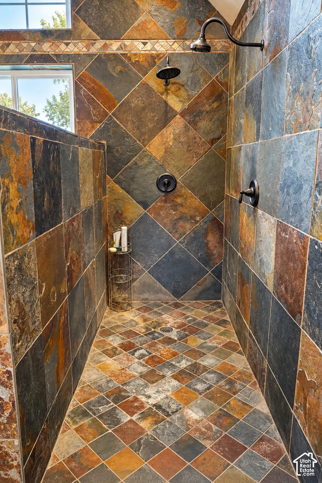 Walk-in shower featuring multi-toned tile walls and flooring, a decorative tile border, and dual shower heads