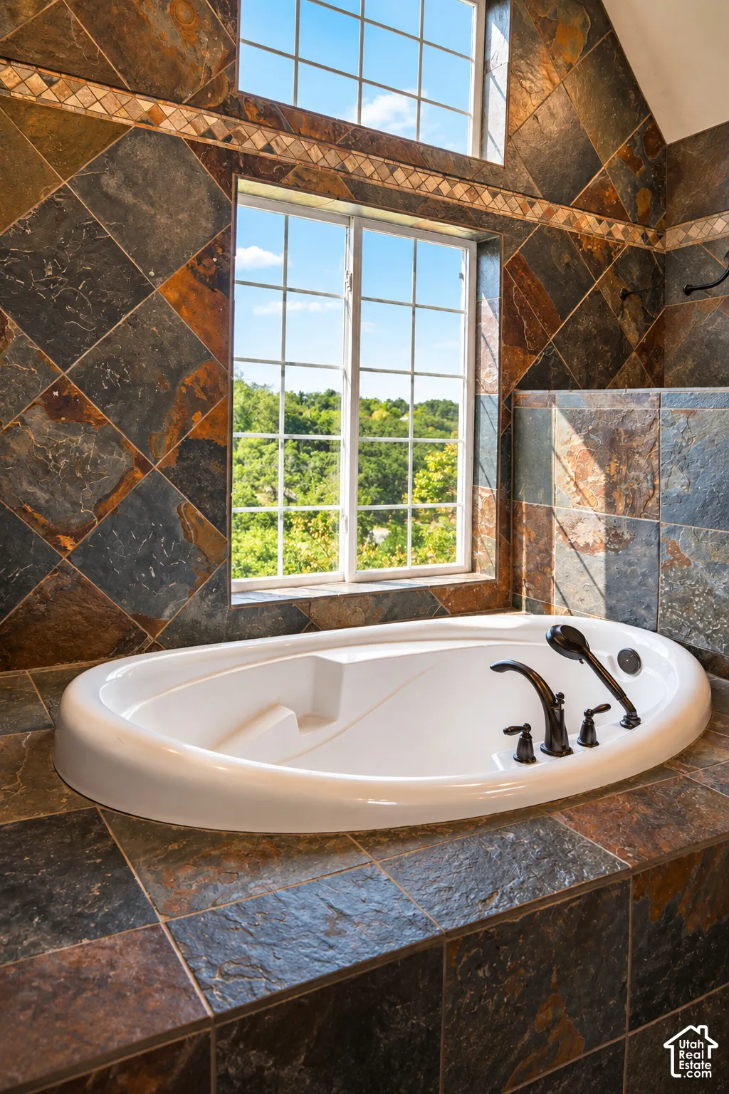 Luxurious soaking tub with dual faucet and integrated armrests, set within a dramatic slate-tile surround featuring warm bronze and charcoal tones in a diagonal pattern