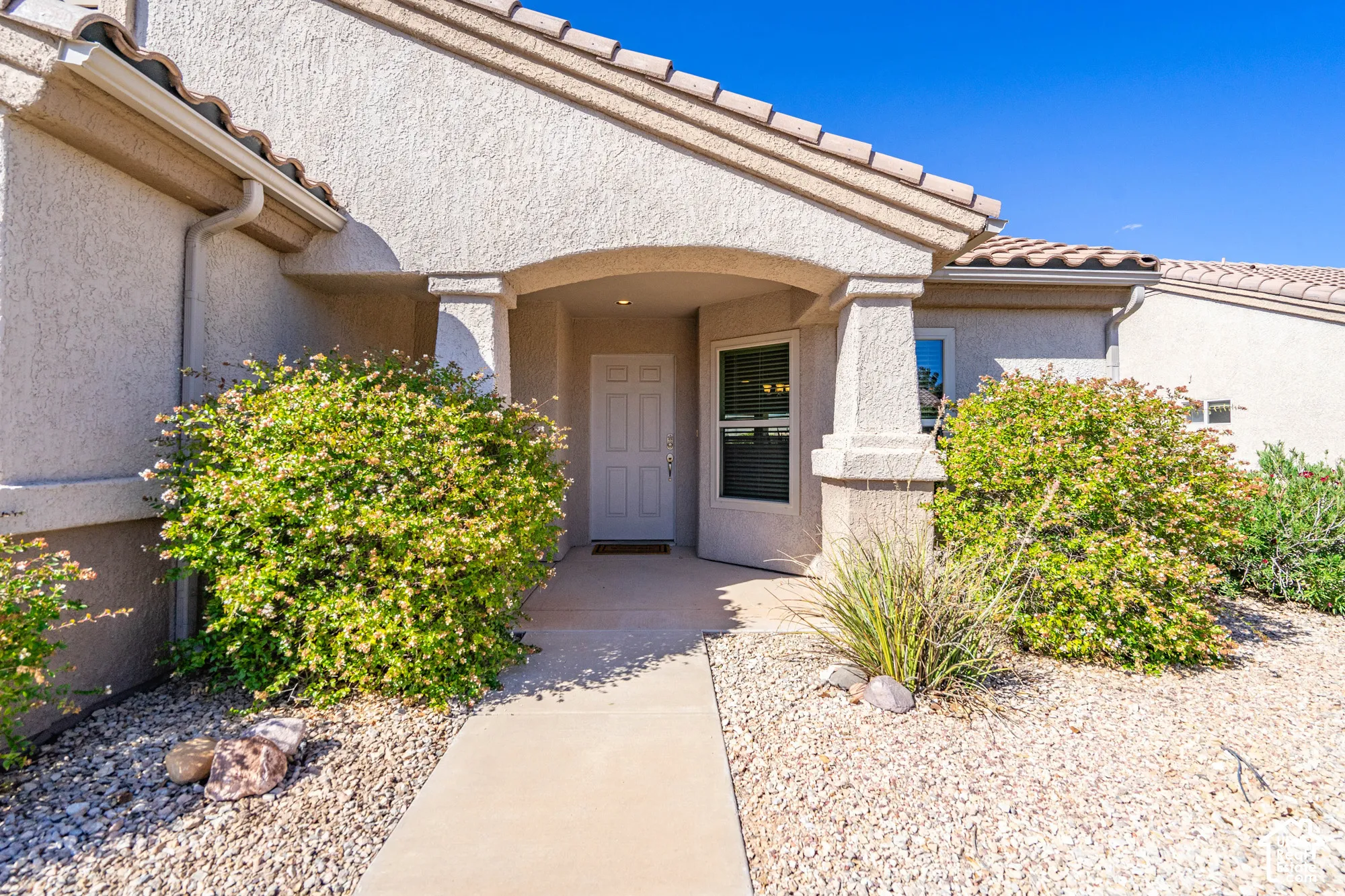 Stucco exterior with a tiled roof, covered entryway, and mature landscaping featuring shrubs and gravel