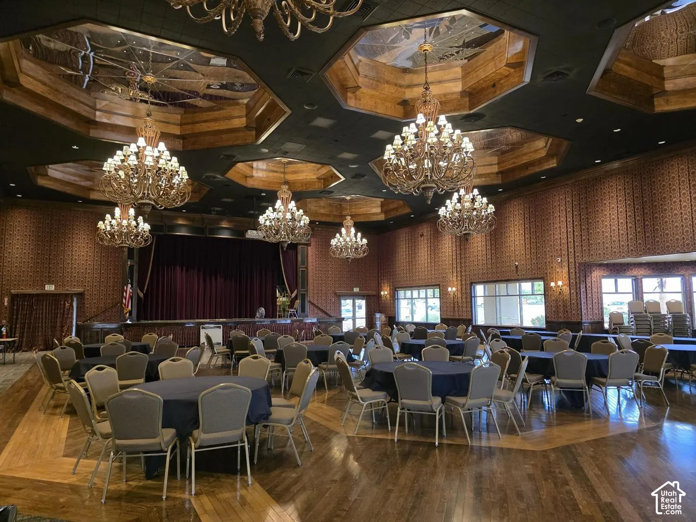 Grand ballroom featuring ornate chandeliers, recessed octagonal ceiling panels, a raised stage with velvet curtains, large windows, and wood-finish flooring