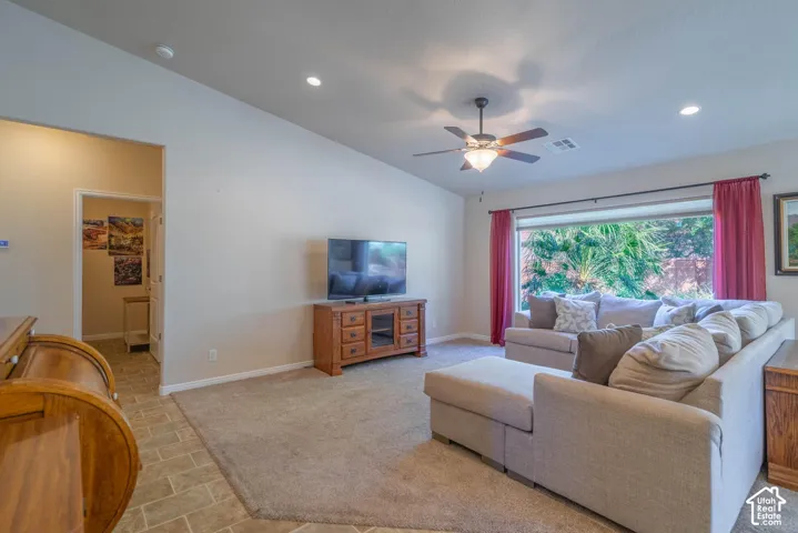 Spacious living room featuring neutral carpeting, a ceiling fan with integrated lighting, recessed ceiling lights, and large windows with pink drapery framing natural light