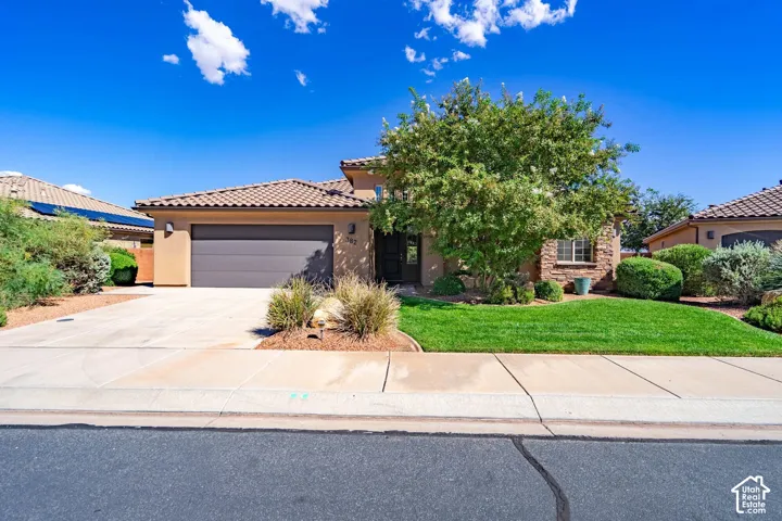 Welcoming front elevation featuring a tan stucco exterior with clay tile roofing, a three-car garage with paneled door, and a professionally landscaped front yard with mature trees, ornamental plantings, and manicured lawn bordered by concrete hardscaping
