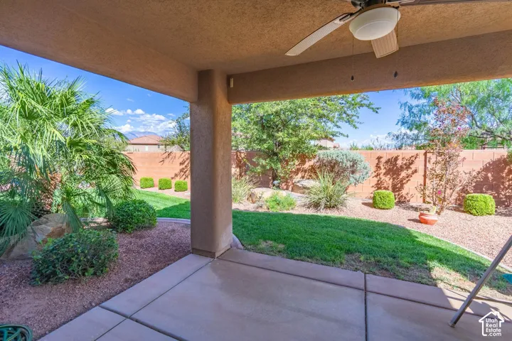 Covered patio with ceiling fan overlooking expansive backyard featuring manicured lawn, mature landscaping with flowering shrubs, decorative rock beds, and brick perimeter fencing