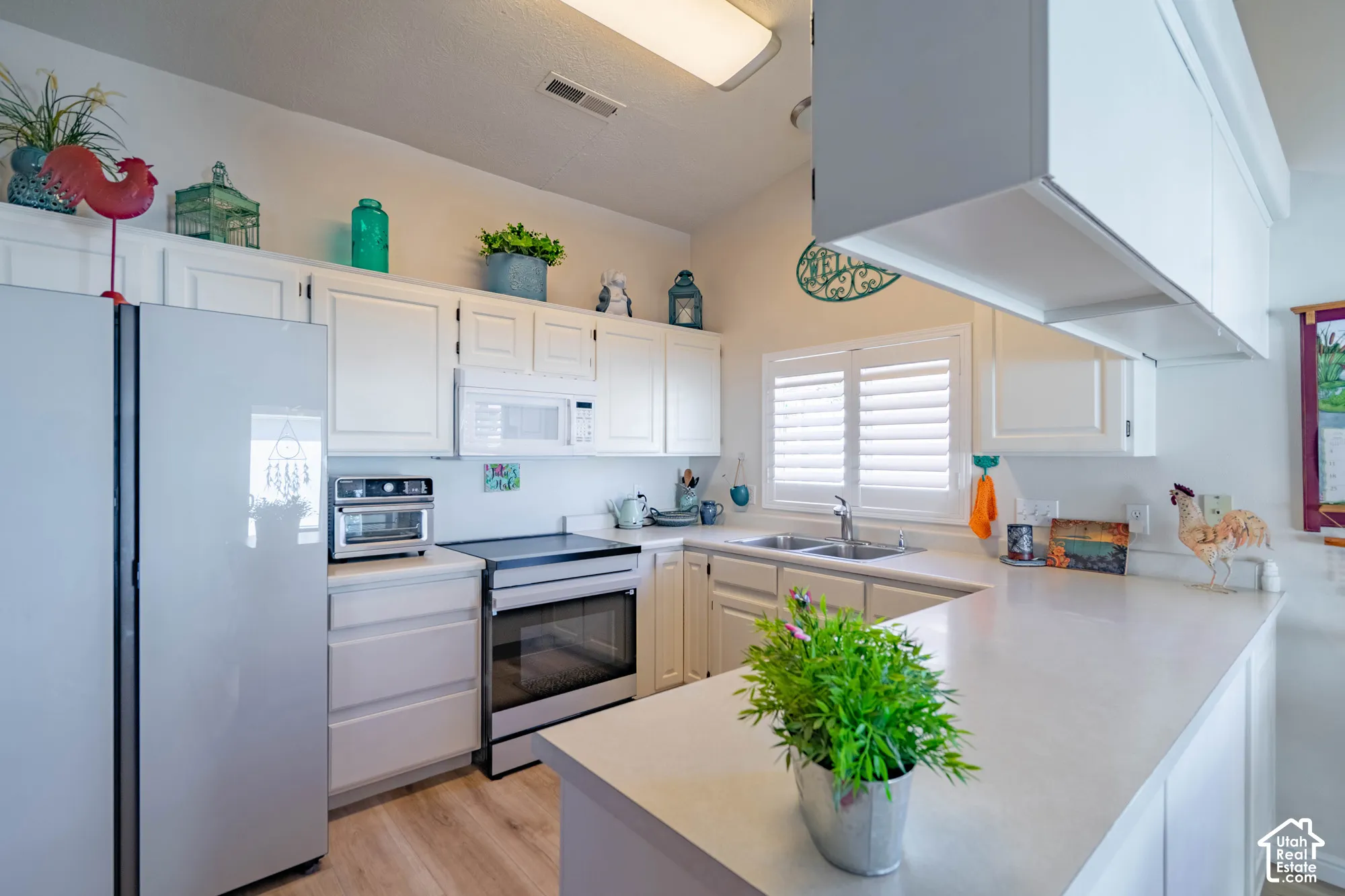 White cabinetry, light-toned countertops, window with plantation shutters, stainless steel appliances, and wood-finish flooring