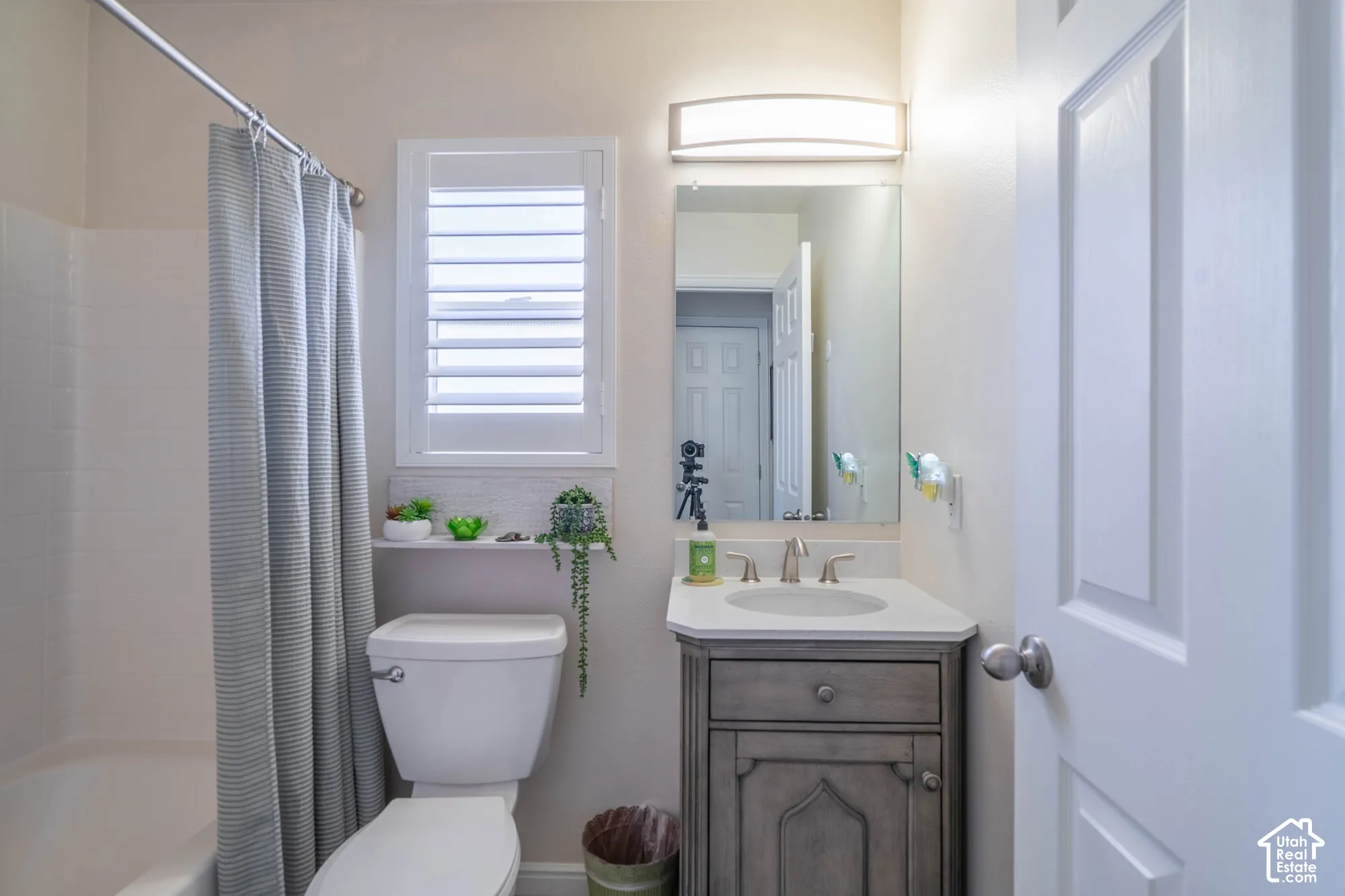 White shaker door, built-in vanity with single basin sink, wall-mounted mirror, overhead vanity lighting, and a white window with louvered shutters