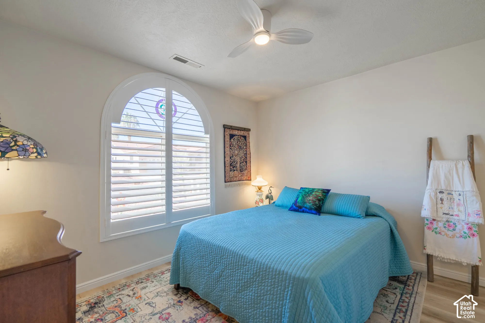 Arched window with white plantation shutters, paddle ceiling fan, and wood-finish flooring