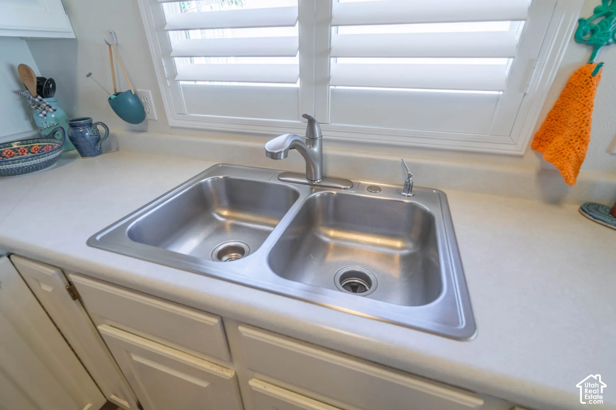 Double basin stainless steel sink with a chrome faucet, set into a light-toned countertop, complemented by white cabinetry and a window with white louvered shutters