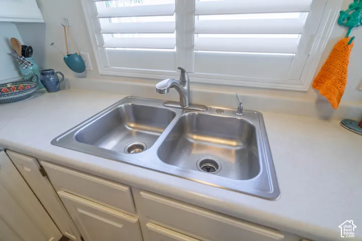 Double basin stainless steel sink with a chrome faucet, set into a light-toned countertop, complemented by white cabinetry and a window with white louvered shutters