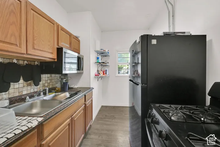 Galley kitchen featuring wood-finish cabinetry, stainless steel countertops and sink, tile backsplash, and gas range