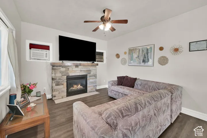 Bright living room featuring a wood-finish plank floor, gas fireplace with stone surround and built-in shelving, wall-mounted flat-screen television, ceiling fan with integrated lighting, and large window with natural light
