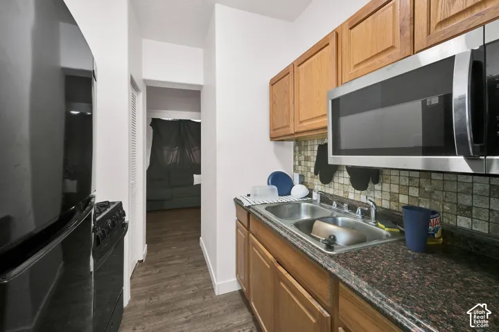 Galley kitchen featuring wood-finish cabinetry, granite-style countertops, stainless steel appliances including a microwave and range, a stainless steel sink, tile backsplash, and wood-finish flooring throughout