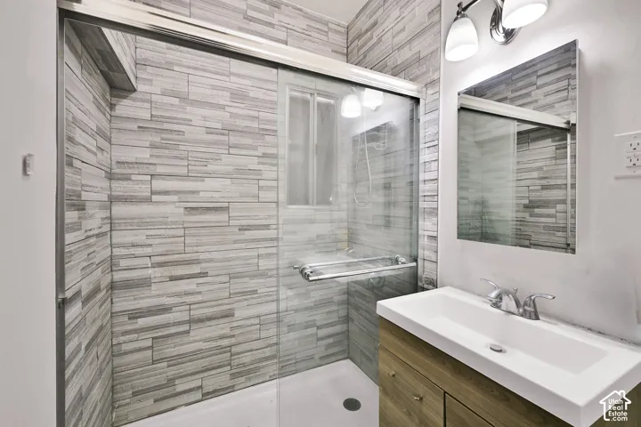 Modern bathroom featuring a linear tile accent wall in gray and white tones, frameless glass shower enclosure with chrome hardware, white rectangular vessel sink, wood-finish vanity with integrated storage, and recessed ceiling lighting
