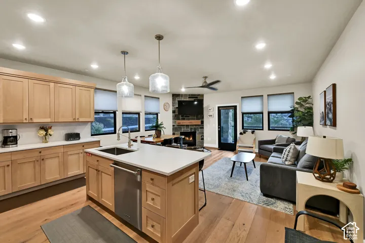 Open-concept living space with wood-finish flooring, a kitchen island with integrated sink, recessed lighting, a stone-clad fireplace, and multiple windows