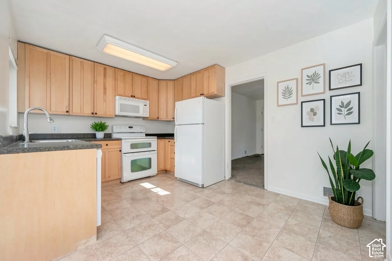 Kitchen featuring neutral tile flooring, light wood-finish cabinetry, dark countertops, and white appliances
