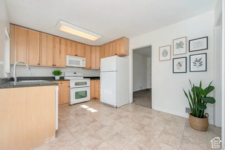 Kitchen featuring neutral tile flooring, light wood-finish cabinetry, dark countertops, and white appliances