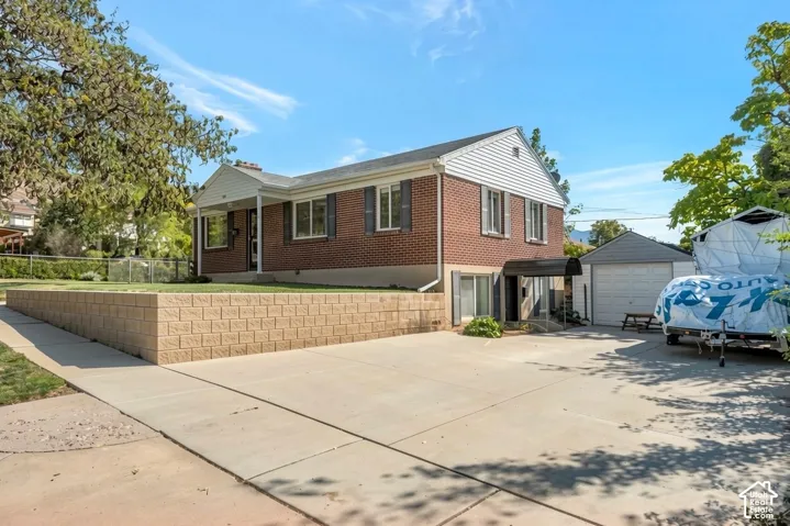 Brick and siding exterior, expansive concrete driveway, detached garage, covered patio entrance, and a terraced retaining wall