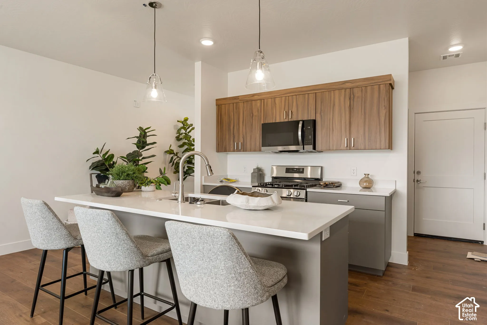 Modern kitchen and dining island featuring warm wood-finish cabinetry, stainless steel appliances, quartz countertops, pendant lighting, and upholstered seating for three