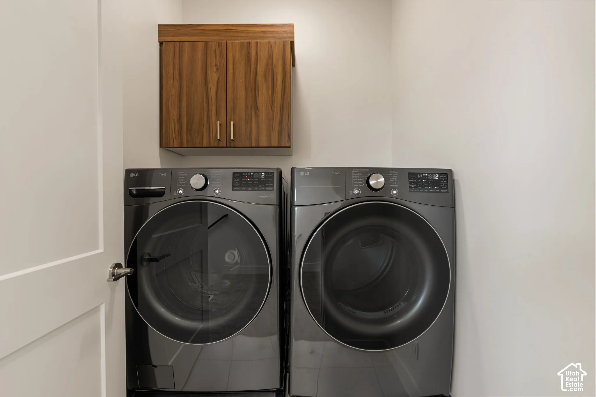 Dedicated laundry room featuring side-by-side front-load washer and dryer with dark gray finish, wooden cabinetry with dual handles above for storage, and neutral walls providing a clean, functional utility space