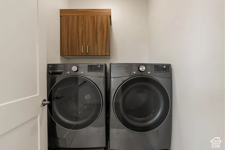 Dedicated laundry room featuring side-by-side front-load washer and dryer with dark gray finish, wooden cabinetry with dual handles above for storage, and neutral walls providing a clean, functional utility space