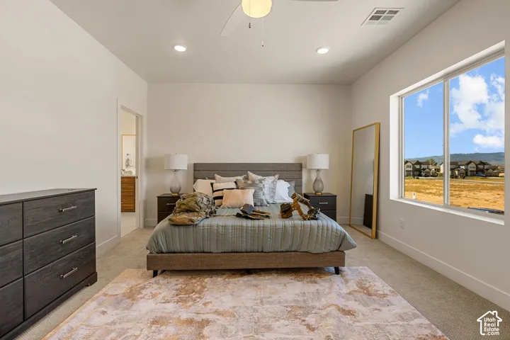 Spacious primary bedroom featuring a wood-finish platform bed frame, dual nightstands with table lamps, neutral wall-to-wall carpeting with an area rug, recessed ceiling lighting, and expansive windows with countryside vistas