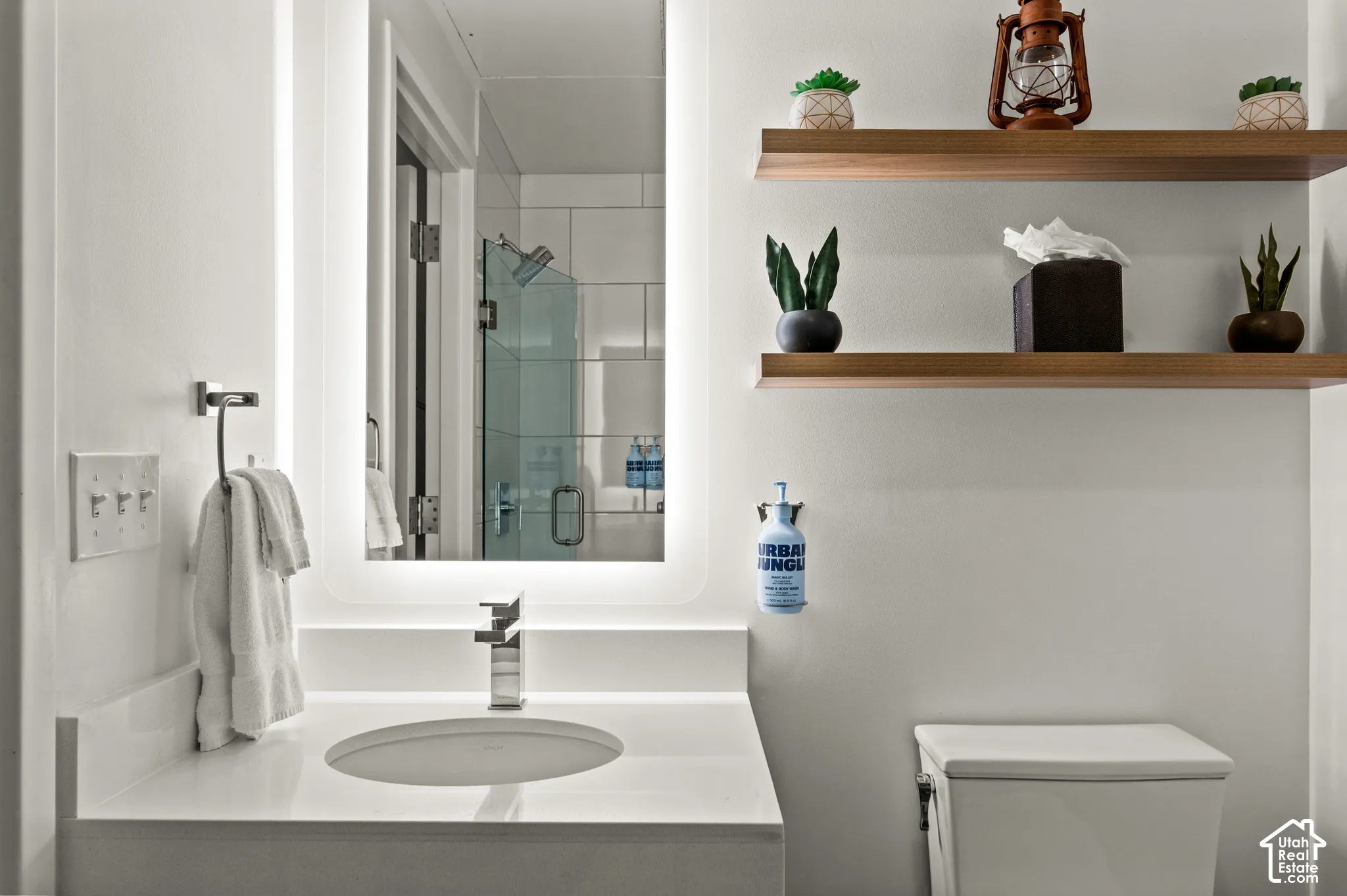 Floating wood shelves, illuminated vanity mirror, integrated sink, wall-mounted faucet, and a frameless glass shower enclosure