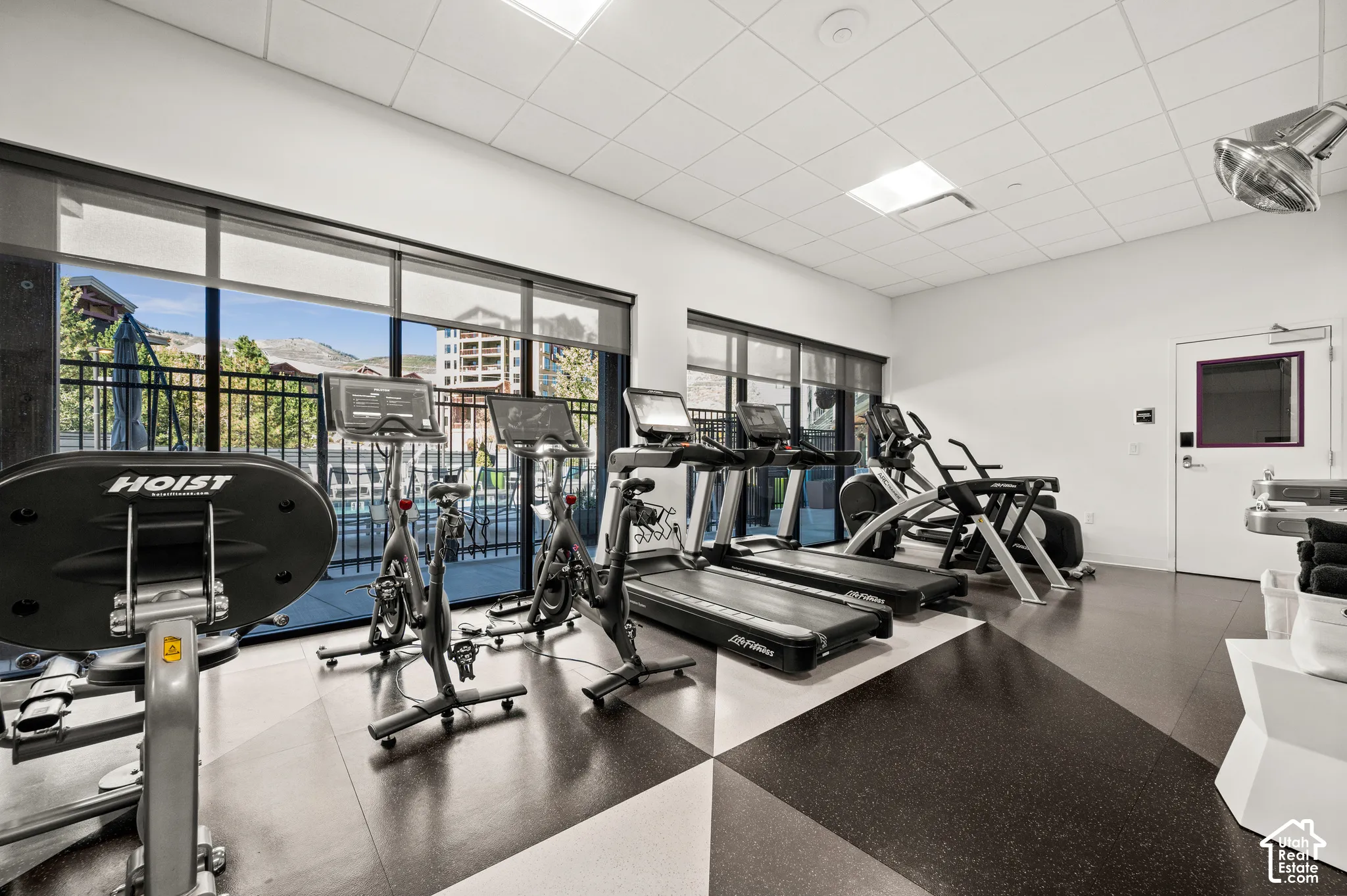 Fitness area with large windows, black and white geometric flooring, white walls, and a suspended ceiling with recessed lighting