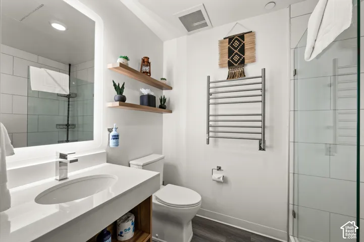 Modern bathroom featuring a vanity with an integrated sink, backlit mirror, glass-enclosed shower, wall-mounted towel warmer, and floating wood shelves